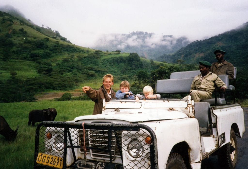 One white adult man (James Meyer) and two young boys (Peter and Jamie) standing behind white truck occupied by two Black adult men in uniform. Two buffalo stand behind them, and there is an expanse of green hills, trees and heavy mist in the background